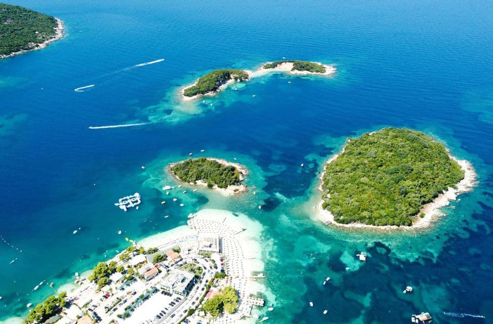 An aerial view of a coastline with small, green islands, a busy beach with umbrellas, and boats on the turquoise sea.