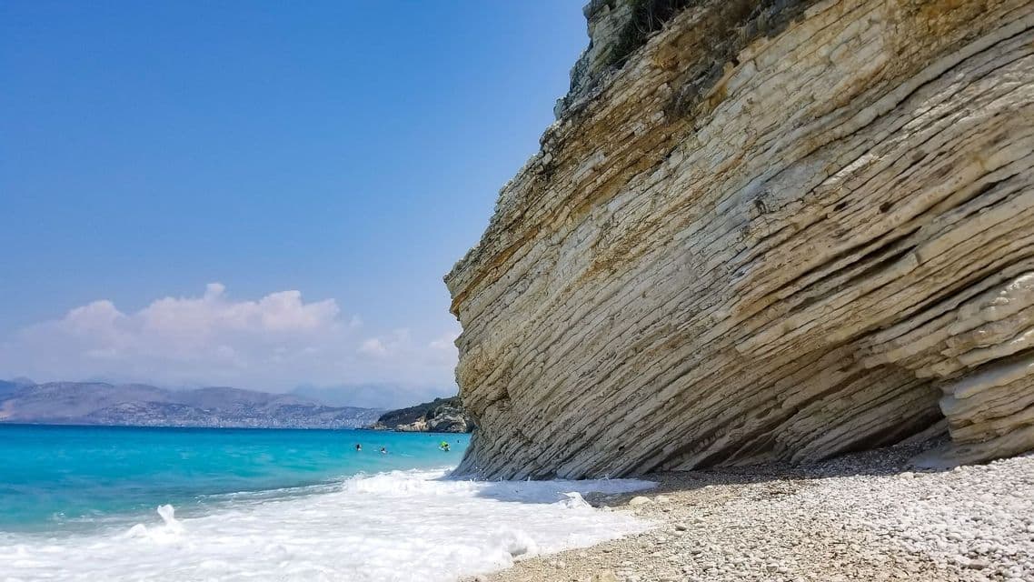 A large, layered rock cliff on a pebble beach with turquoise water and gentle waves under a clear blue sky.