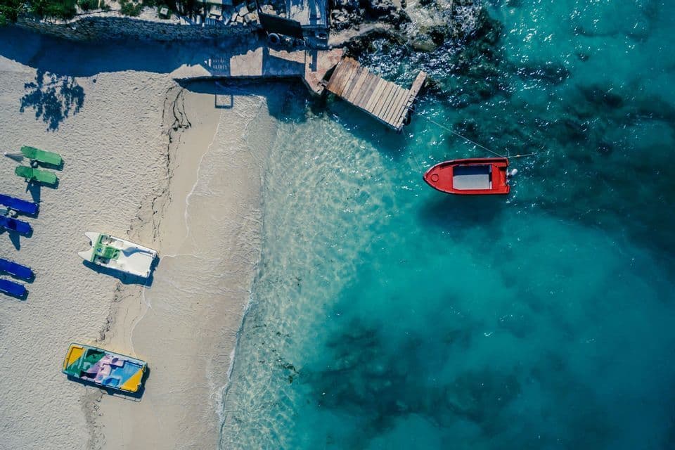An aerial view of a sandy beach with boats on the shore and a red boat moored to a dock in clear turquoise water.