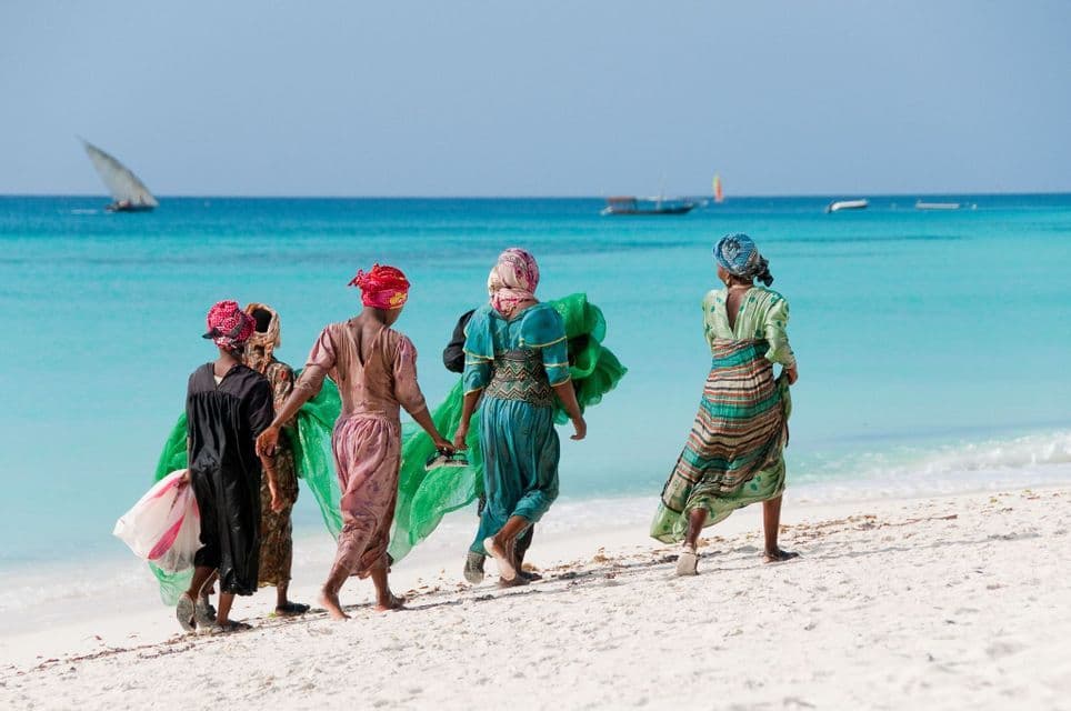 Un groupe de femmes vêtues de tenues traditionnelles colorées marche le long d'une plage de sable blanc au bord d'un océan turquoise, avec des bateaux en arrière-plan.