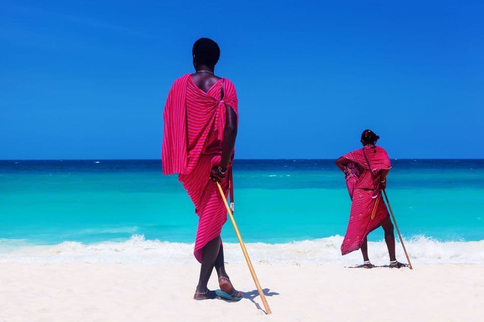 Dos personas con vestimentas tradicionales rosadas están de pie con bastones en una playa de arena blanca, mirando el océano turquesa.