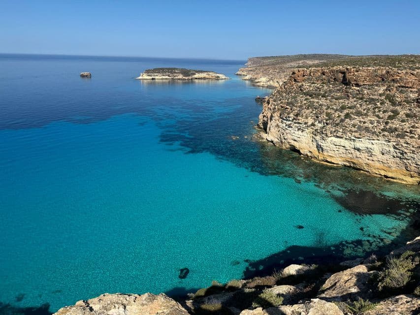 Una vista dall'alto di una costa rocciosa con una baia calma di acqua turchese limpida sotto un cielo azzurro brillante.