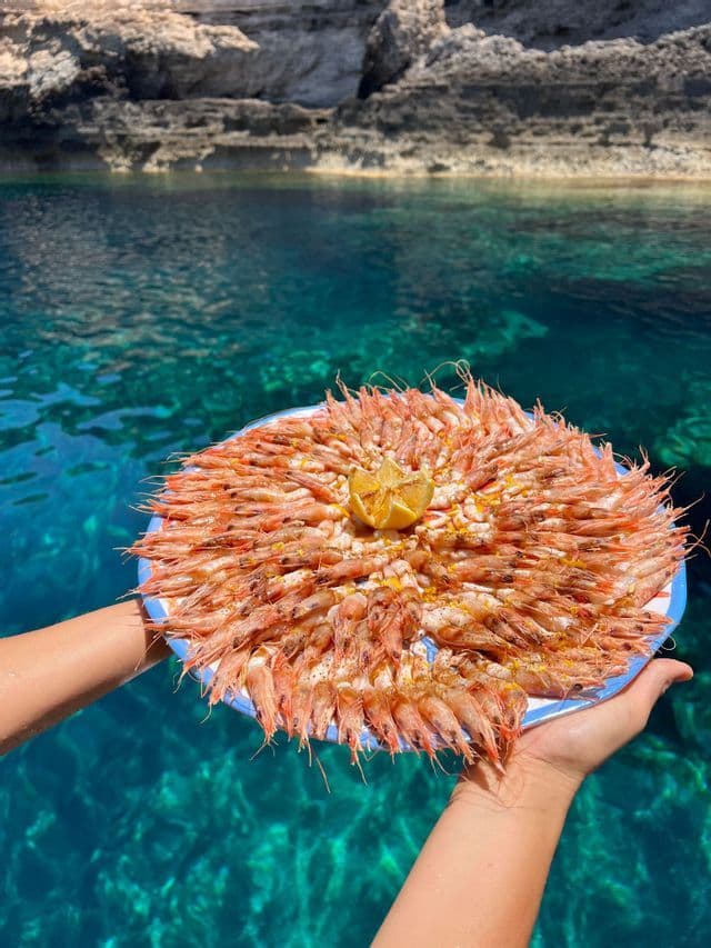 Una persona tiene un grande piatto di gamberi cotti con uno spicchio di limone su acqua cristallina turchese vicino a una costa rocciosa.