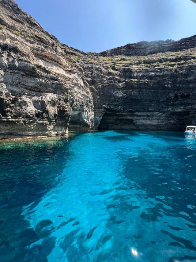 La luce del sole illumina l'acqua limpida e turchese di una cala rocciosa, dove una piccola barca galleggia vicino a una grotta marina.
