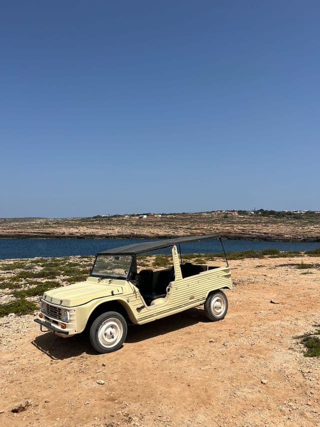 Una decappottabile d'epoca color crema parcheggiata su una strada sterrata su una scogliera a picco sul mare.