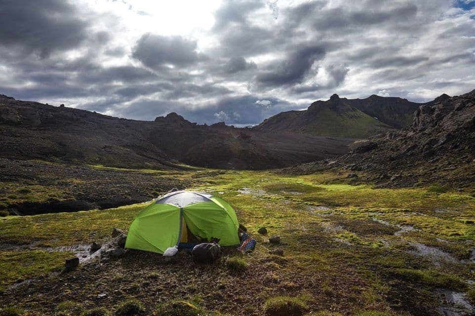 A green camping tent is pitched in a vast, mossy valley surrounded by dark, rocky mountains under a dramatic, cloudy sky.