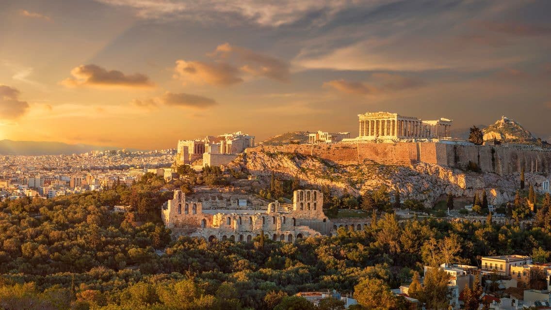 Una vista panorámica de ruinas antiguas en una colina, rodeadas de árboles y con vistas a una vasta ciudad durante un atardecer dorado.