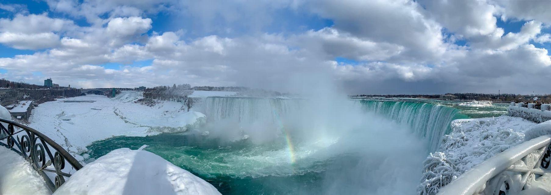 A panoramic view of a wide, powerful waterfall in winter, with snow and ice covering the landscape and a rainbow in the mist.