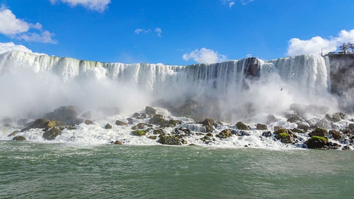 Une large et puissante cascade déferle d'une falaise sur de gros rochers en contrebas, créant un épais brouillard sous un ciel bleu.