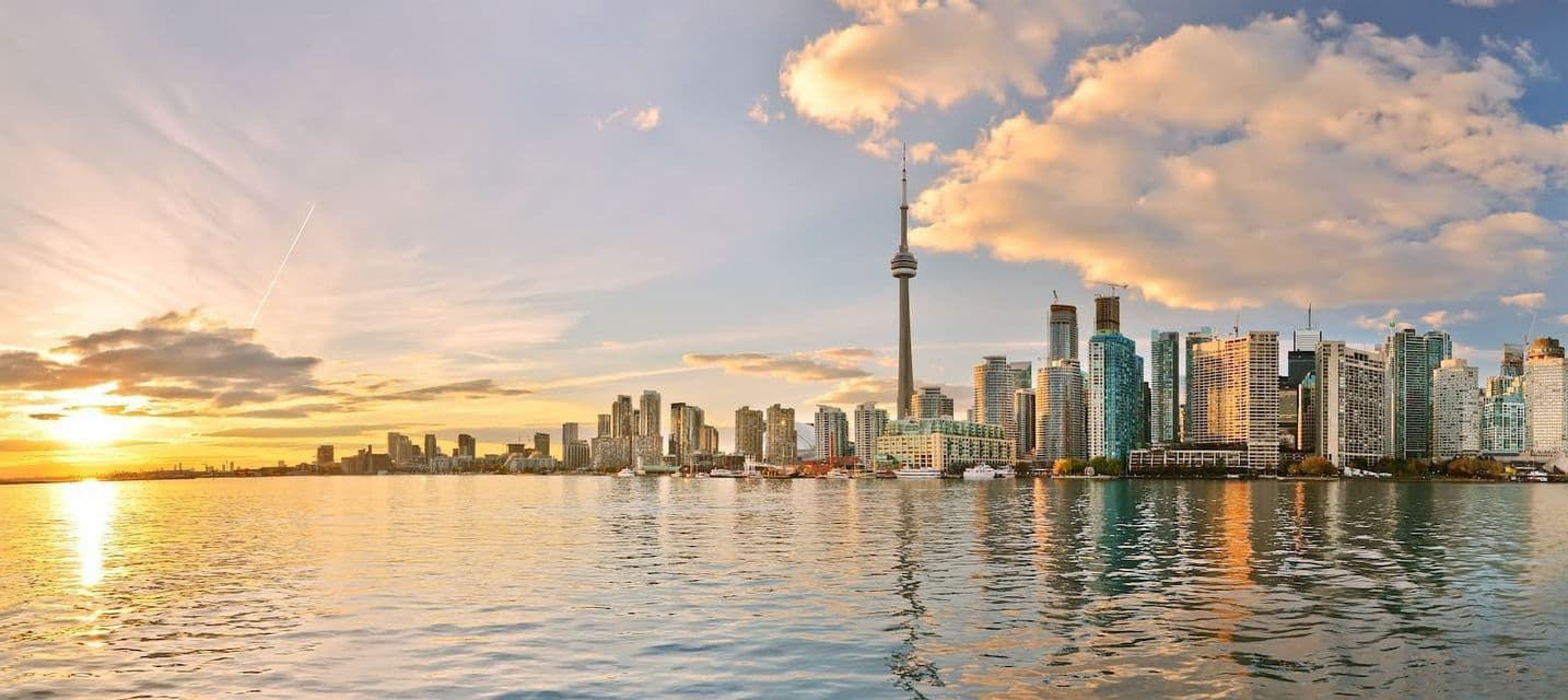 A panoramic city skyline with a tall tower and skyscrapers reflecting on the water at sunset under a partly cloudy sky.