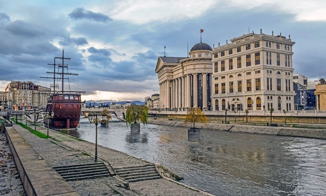Un grande edificio neoclassico e un grande galeone in legno costeggiano le rive di un fiume cittadino sotto un cielo nuvoloso.