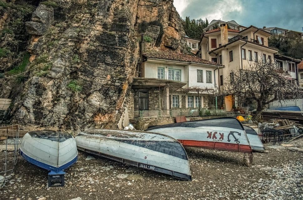 Diverse piccole imbarcazioni capovolte sulla spiaggia di ciottoli ai piedi di una scogliera rocciosa con case incastonate nella collina.
