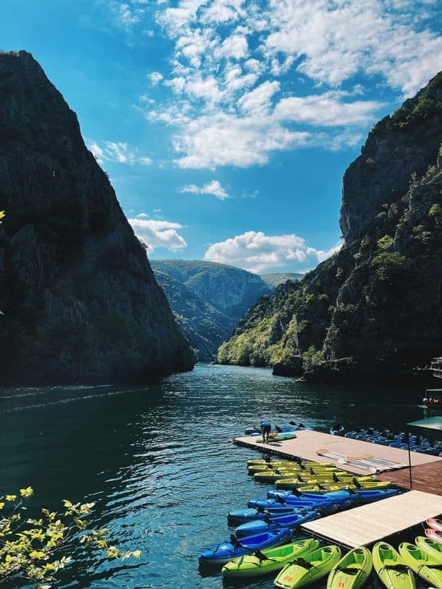 Un pontile di legno galleggiante con molti kayak colorati si trova su un fiume che serpeggia attraverso un ripido canyon roccioso sotto un cielo blu.