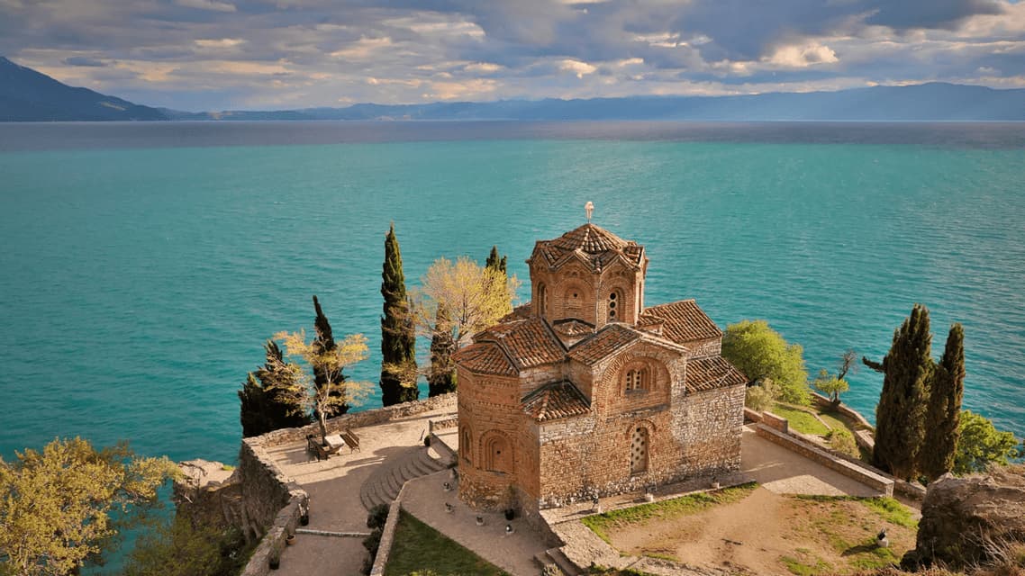 Un'antica chiesa in pietra con tetto in tegole sorge su una scogliera che domina un vasto lago turchese sotto un cielo nuvoloso.