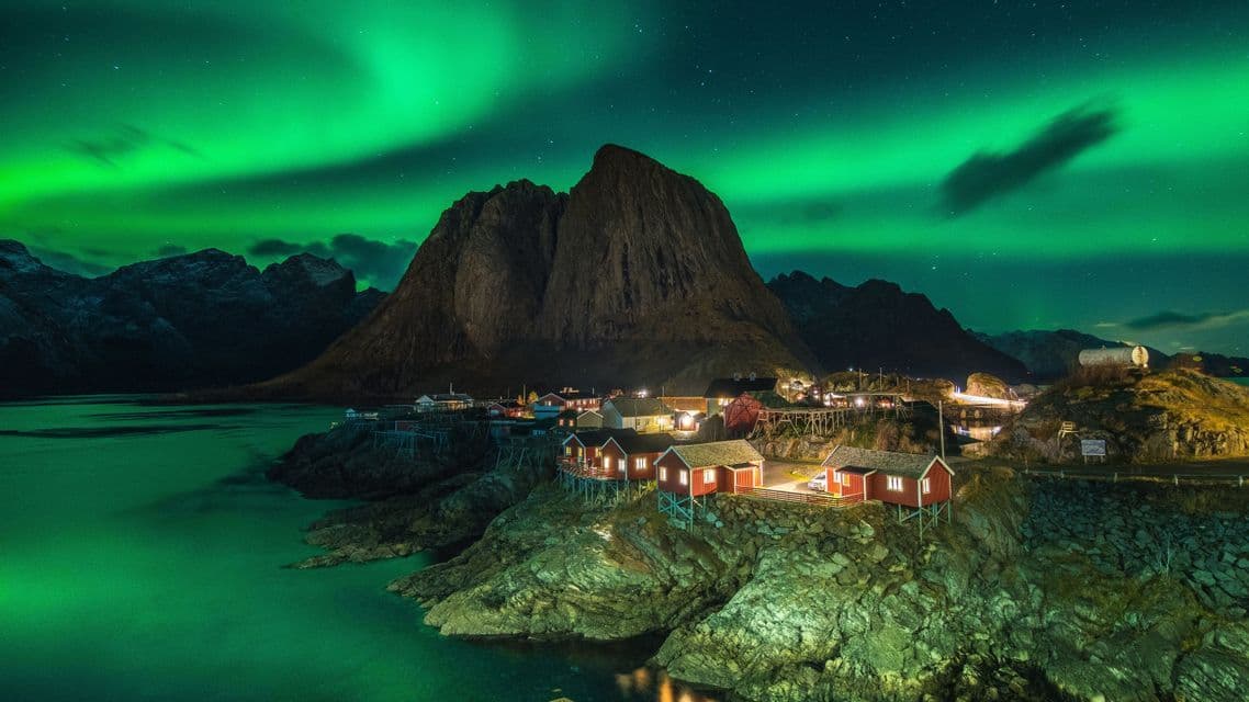 La aurora boreal verde brilla en el cielo nocturno sobre un pueblo costero iluminado con casas rojas al pie de una montaña.