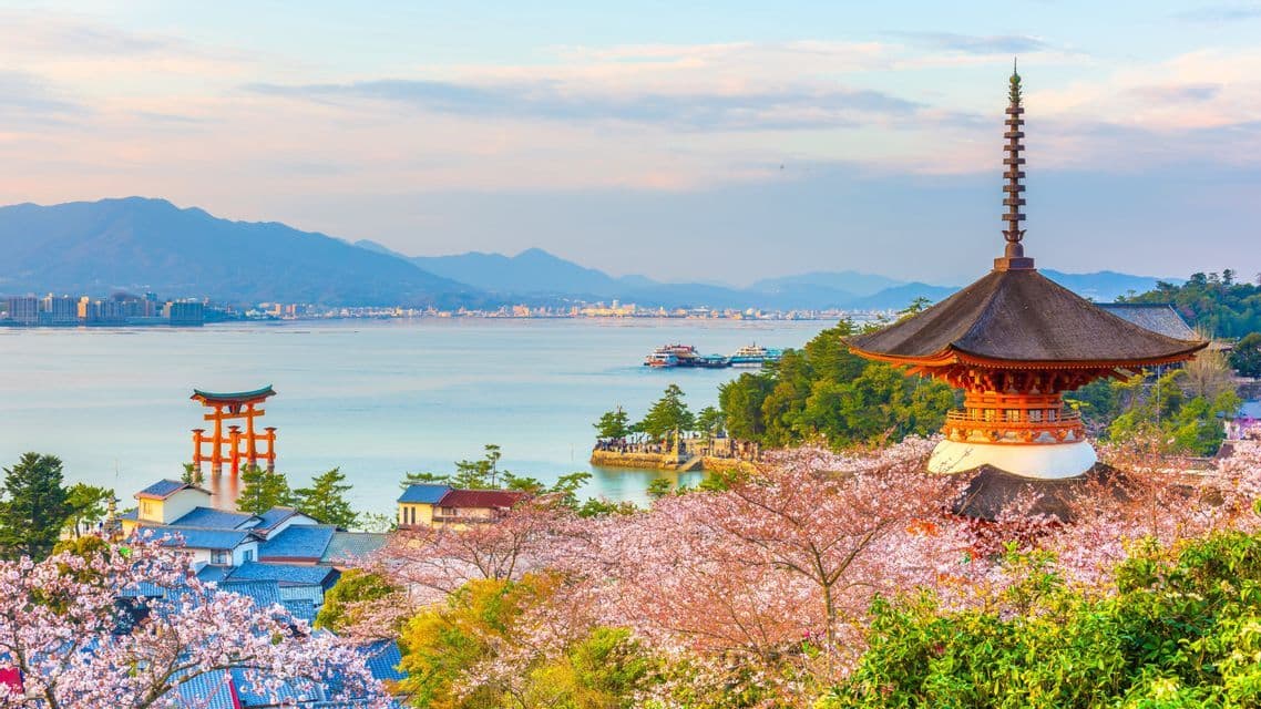 Una pagoda giapponese e fiori di ciliegio si affacciano su una baia con un cancello torii rosso e montagne lontane.