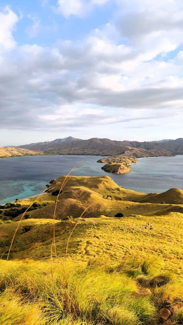 Eine WeRoad-Gruppe steht auf einem sonnenbeschienenen, goldenen Hügel und blickt auf eine Bucht mit kargen Inseln unter einem teils bewölkten Himmel.