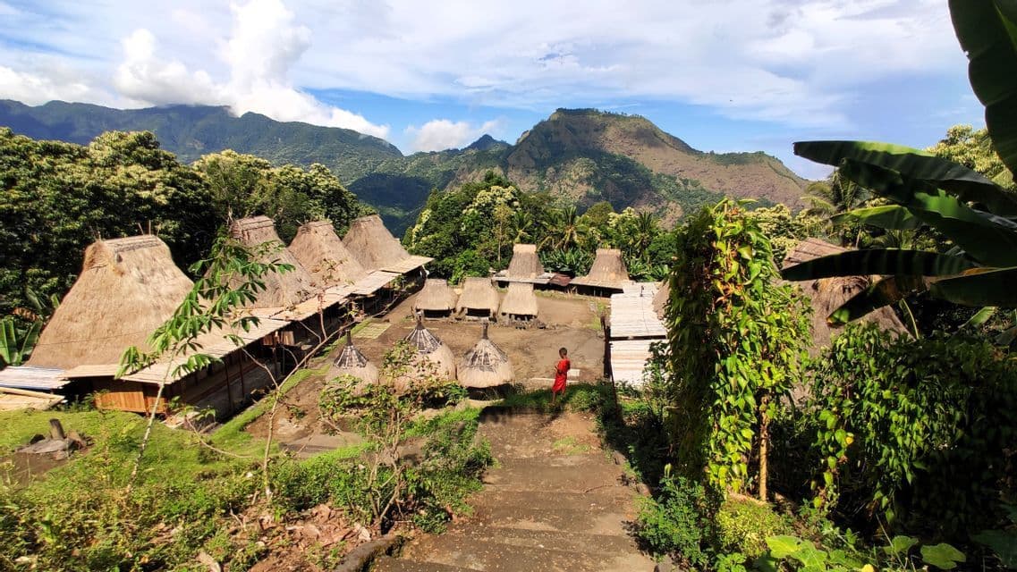 Ein Dorf mit kegelförmigen strohgedeckten Hütten, eingebettet in einem üppig grünen Bergtal, mit einem Kind in Rot auf einem Steinpfad.