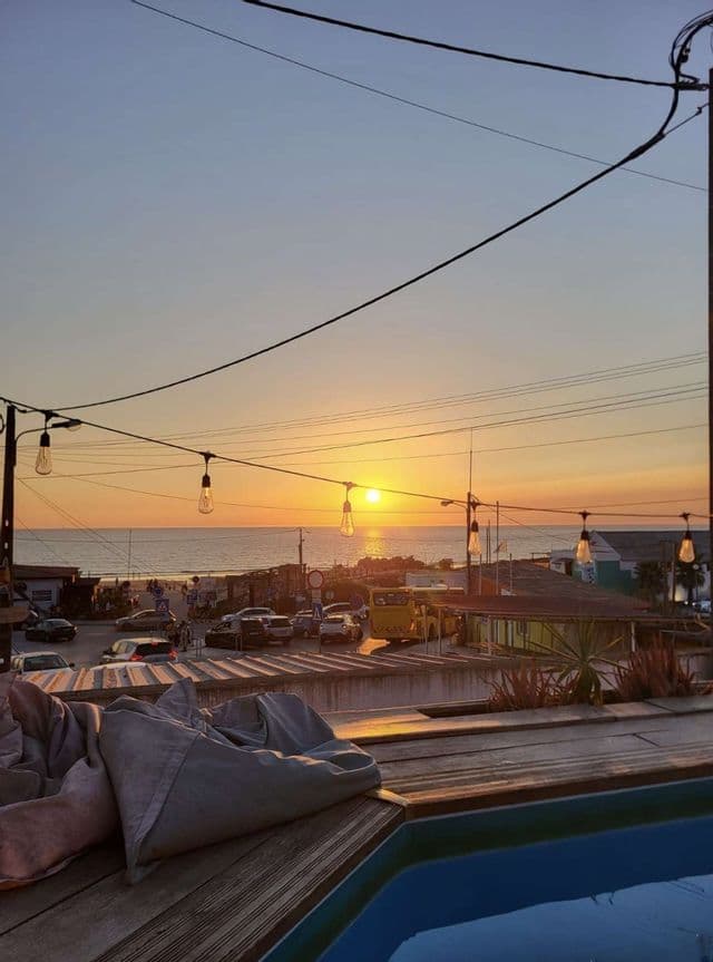 Luci decorative pendono su una terrazza in legno con piscina, affacciata su una cittadina di mare al tramonto sull'oceano.