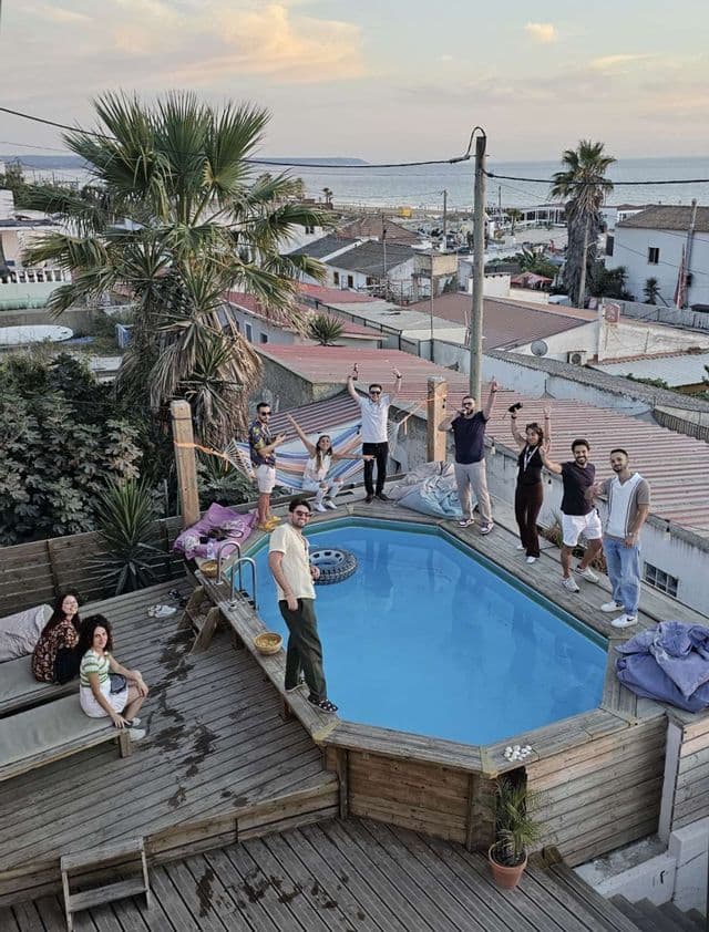 Un viaggio di gruppo WeRoad di amici che posano felicemente su una terrazza panoramica in legno intorno a una piscina, con una città costiera sullo sfondo.