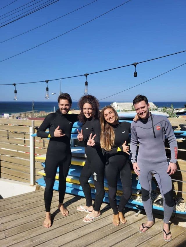 Un viaggio di gruppo WeRoad in mute da sub che posano insieme su una terrazza di legno con tavole da surf e l'oceano sullo sfondo.