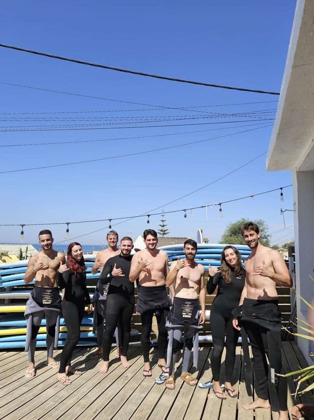 Un gruppo WeRoad in posa in mute su una piattaforma di legno davanti a una rastrelliera di tavole da surf.