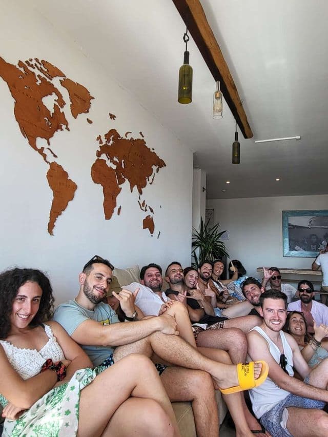 Un gruppo WeRoad in viaggio è seduto su un divano, sorridendo per una foto davanti a una mappa del mondo in legno.