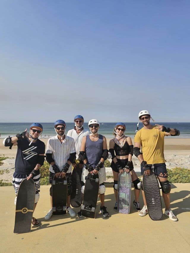 Un viaggio di gruppo WeRoad con persone che indossano caschi e protezioni in posa con i loro skateboard su una spiaggia in una giornata di sole.