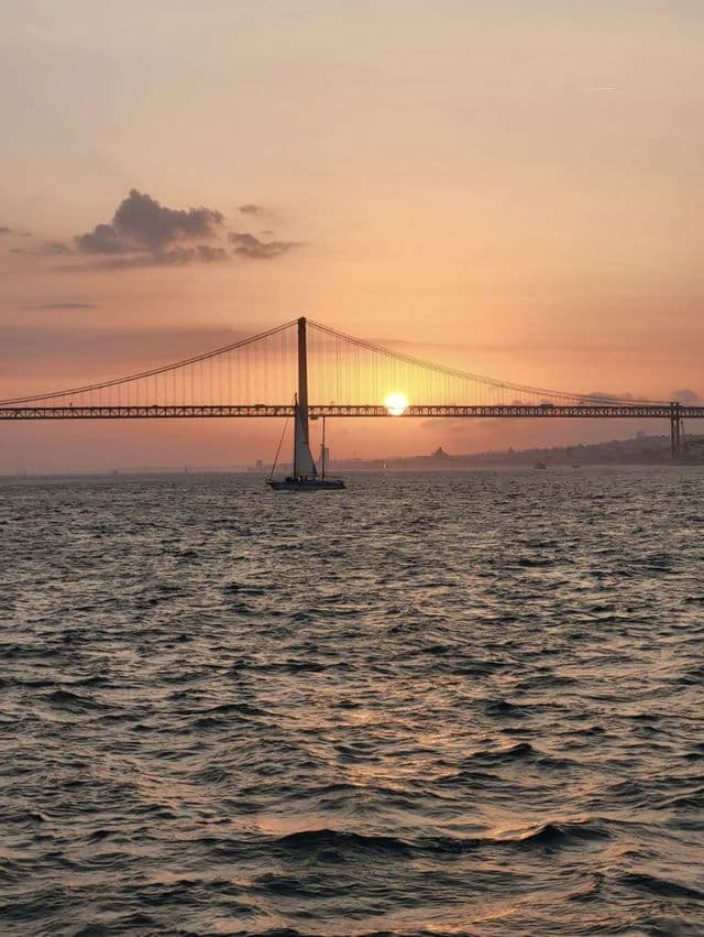 Una barca a vela scivola sulle acque agitate davanti a un grande ponte sospeso mentre il sole tramonta alle sue spalle.