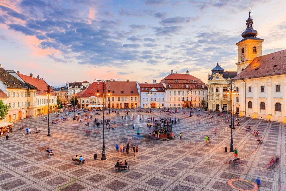 Una veduta dall'alto di un'ampia piazza lastricata a ciottoli con motivi, con persone in movimento sfocate tra edifici storici al tramonto.