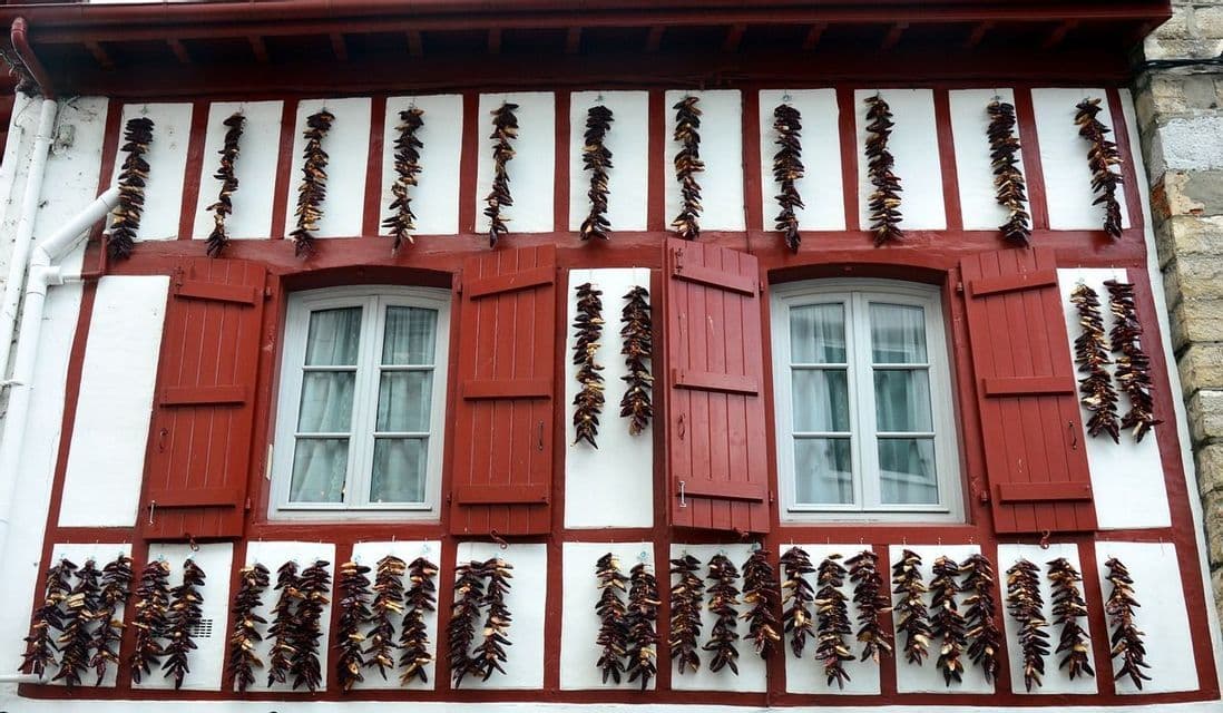 Des guirlandes de poivrons rouges séchés pendent sur la façade d'un bâtiment avec des colombages rouges et des volets rouges assortis.