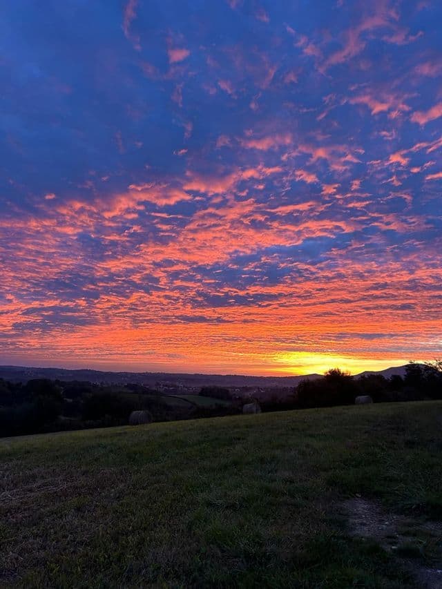 Un coucher de soleil éclatant avec des nuages roses et oranges sur un ciel bleu profond, vu d'une colline herbeuse dominant une vallée.