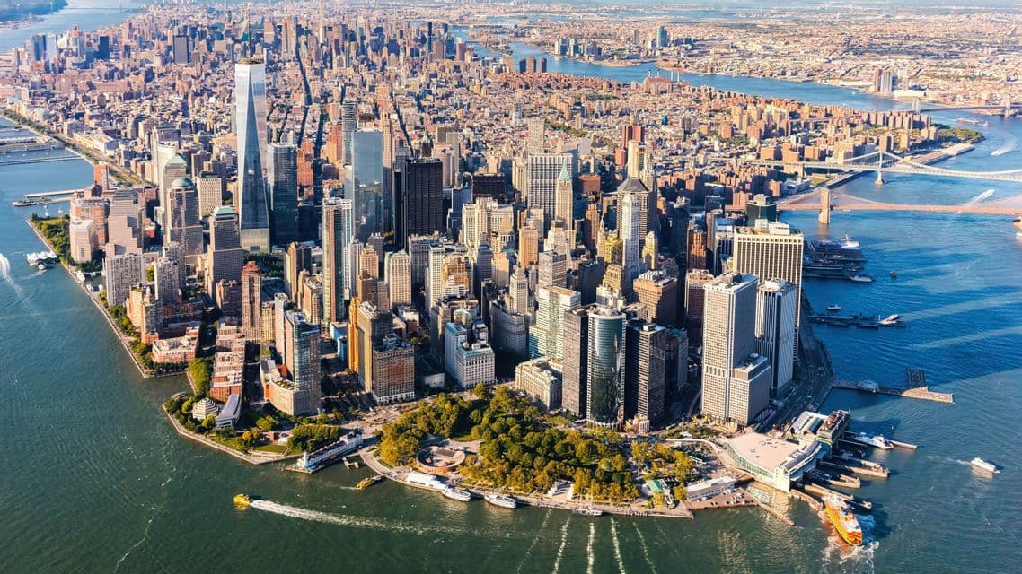 Aerial view of the Lower Manhattan skyline, with dense skyscrapers surrounded by rivers on a sunny day.