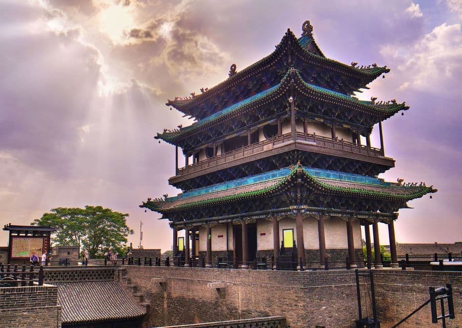 A traditional Chinese pagoda with green-tiled roofs sits atop a stone wall under a sky with clouds and piercing sunbeams.