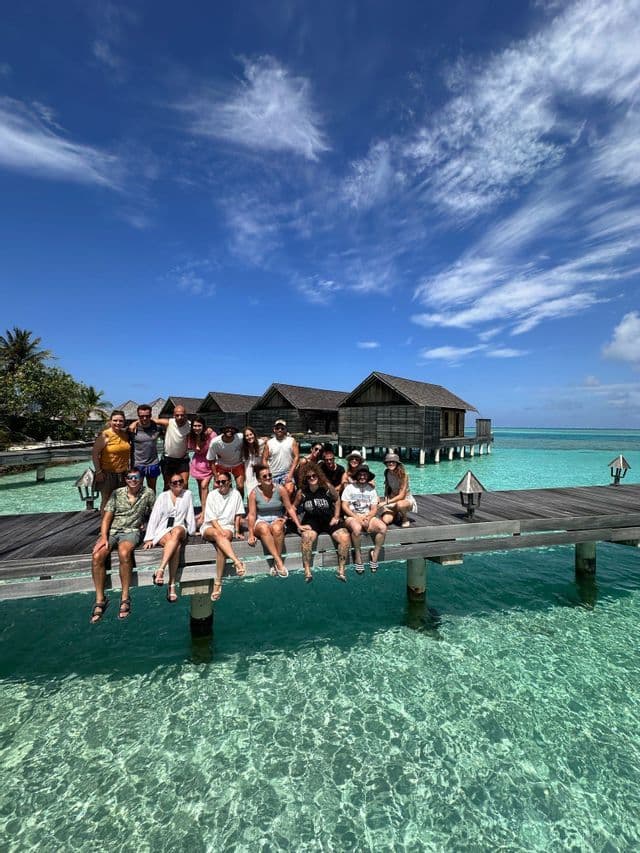 Un viaje en grupo de WeRoad posa en un muelle de madera sobre agua turquesa cristalina, con bungalows sobre el agua al fondo bajo un cielo azul.