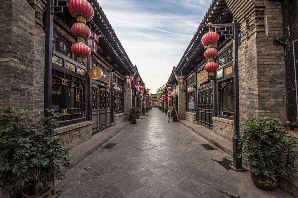 A narrow paved street flanked by traditional brick buildings decorated with red paper lanterns under a cloudy sky.