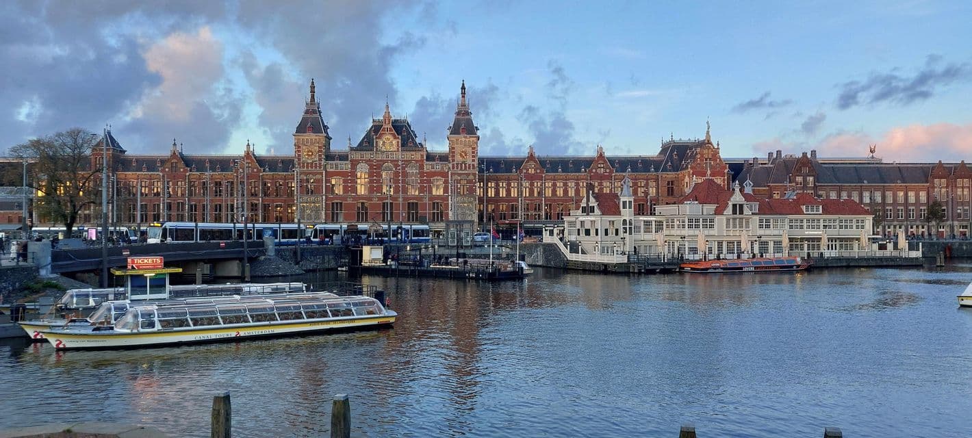 Un gran edificio de ladrillo rojo visto desde el otro lado de un canal, con barcos turísticos y un tranvía en primer plano bajo un cielo nublado.