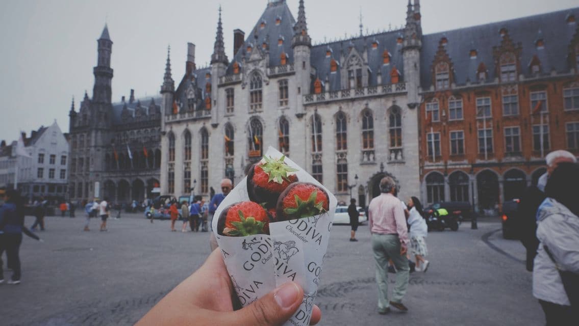 Una mano sostiene un cucurucho de papel de fresas cubiertas de chocolate Godiva en una plaza histórica de una ciudad europea.