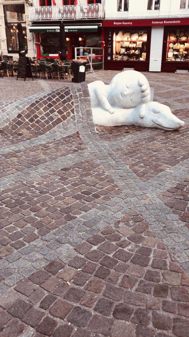 Una escultura de piedra blanca de un niño durmiendo sobre un perro, parcialmente cubierto por una manta de adoquines que se funde con la plaza.