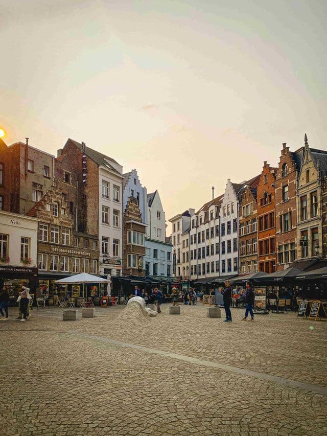 Una plaza adoquinada con gente paseando entre cafés, rodeada de edificios históricos con tejados a dos aguas bajo un cálido atardecer.