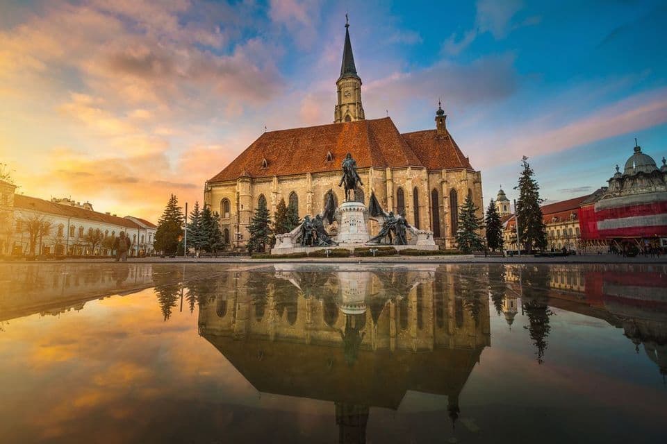Una grande chiesa di pietra e un monumento riflessi in una grande pozzanghera in una piazza cittadina sotto un cielo al tramonto colorato.