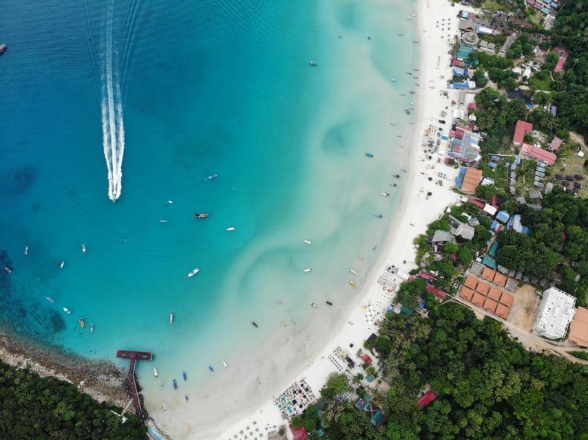 Luftaufnahme von einem tropischen Strand mit einem Motorboot, das eine Bugwelle im türkisfarbenen Wasser zieht, und am Ufer vertäuten Booten.