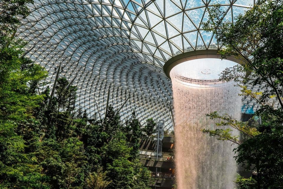 Ein Indoor-Wasserfall strömt aus einem Oberlicht in einer großen, geometrischen Glaskuppel, umgeben von üppigen grünen Bäumen und Laub.