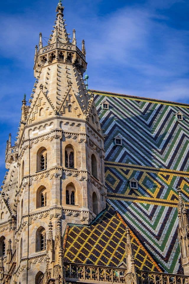 An ornate stone tower of a Gothic cathedral with a colorful, patterned tile roof against a bright blue sky.