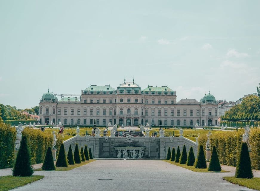 Ein weiter Blick auf einen prächtigen Palast mit grünem Dach, gelegen hinter einem formalen Garten mit Springbrunnen und einem zentralen Weg.