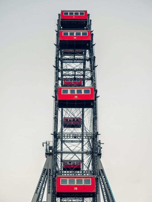 A low-angle shot of a Ferris wheel with a dark metal frame and red, numbered cabins against a plain, overcast sky.
