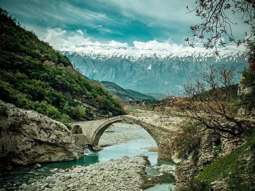 Un ponte ad arco in pietra attraversa un fiume in una valle rocciosa, con imponenti montagne innevate sullo sfondo.