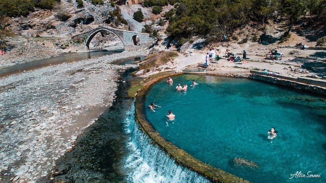 Veduta aerea di un viaggio di gruppo WeRoad che nuota in una piscina termale turchese, con un ponte ad arco in pietra e un fiume sullo sfondo.