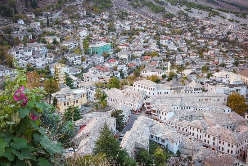 Una vista aerea di una città storica con edifici bianchi e tetti in pietra, incastonata in una collina verde e lussureggiante.