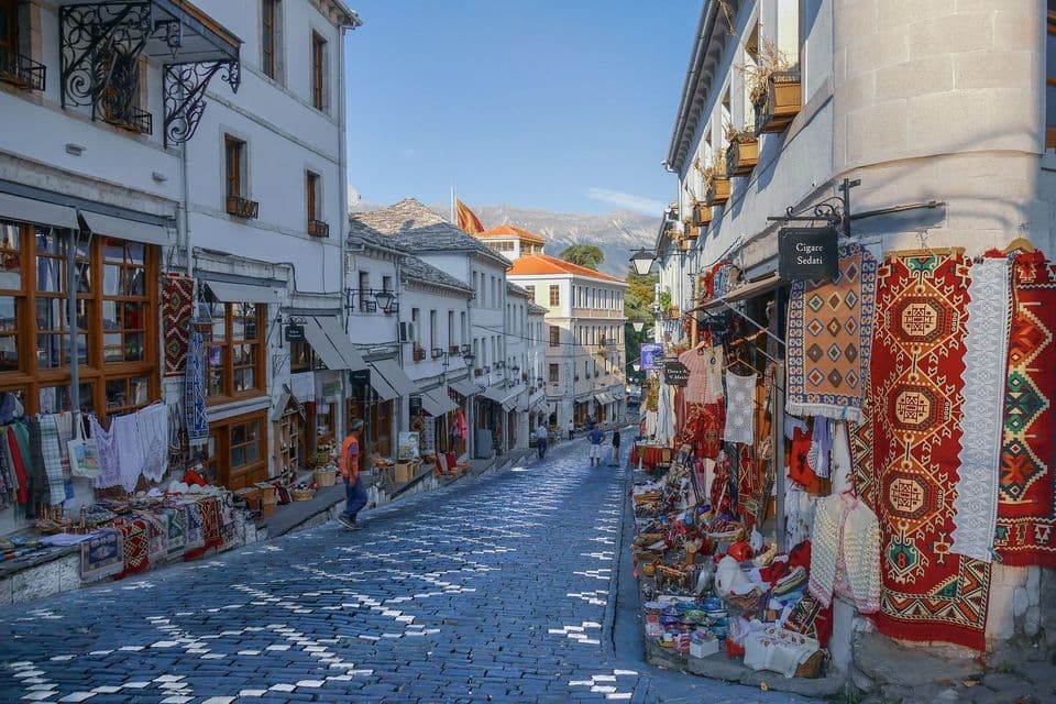 Une rue pavée ornée de motifs, bordée de boutiques exposant des tapis et textiles colorés, avec des montagnes visibles au loin.